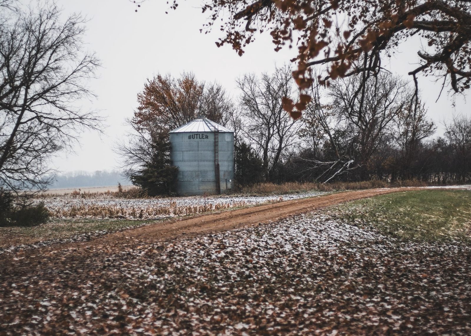 A dirt farm road past a single grain bin and bare trees, first snow on the ground